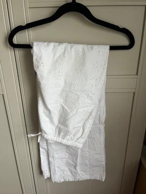 Vita broderade linnebyxor - Säljer ett par vita linnebyxor med broderade blommor och resår i midjan. Byxorna har raka ben och en avslappnad passform, perfekta för en chill och somrig look. Materialet känns lätt och luftigt, och de har snörning i midjan för extra komfort. 