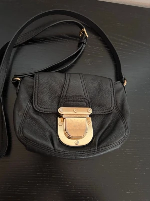 Svart handväska från Michael Kors - Snygg svart handväska i mjukt läder från Michael Kors med justerbar axelrem och guldfärgat metallspänne framtill. Väskan har en rundad form och lockstängning med tydlig logga på spännet. Perfekt för dig som gillar klassisk och stilren design.