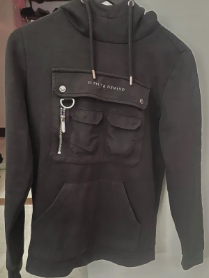 Svart hoodie Supply & Demand XS - Svart hoodie från Supply & Demand med stor ficka framtill, lite slitage på logo texten, två mindre fickor med lock och dragkedjedetalj. Huva med snören och tryckt logga på bröstet. Skön streetwear-stil, perfekt till jeans eller joggers.