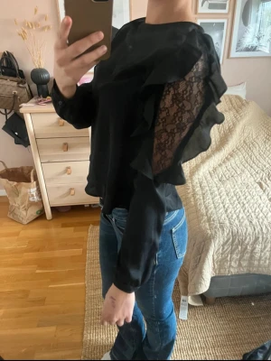 Svart blus med spets och volanger från Zara  - Snygg svart blus med transparent spetsdetalj på ena ärmen och volanger längs axeln och ärmen. Croppad modell med lång ärm och rund halsringning. Perfekt för dig som vill ha något extra till outfiten.