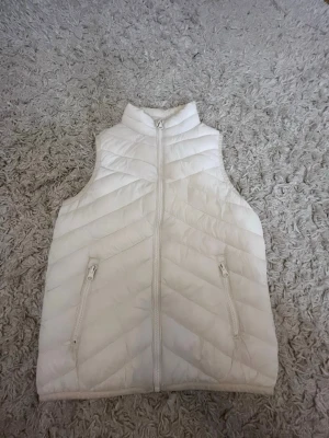 Vit dunväst med dragkedja - Snygg vit dunväst med hög krage och dragkedja framtill. Västen har två fickor med dragkedja och ett quiltat mönster i diagonala ränder. Perfekt att slänga över en hoodie eller tröja för extra värme.