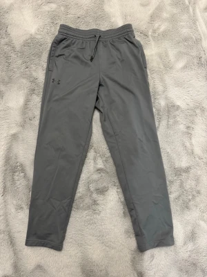 Grå mjukisbyxor från Under Armour - Snygga grå mjukisbyxor från Under Armour med dragsko i midjan och diskret logga på vänster lår. Byxorna har raka ben och är tillverkade i ett mjukt, stretchigt material som känns skönt mot huden. Perfekta för chill eller träning.