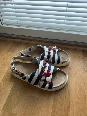 Randiga inuikii sandaler med pärldetaljer - Säljer ett par inuikii sandaler med breda remmar i blåvitt randigt tyg och dekorativa pärlor i rött och vitt. Sandalerna har öppen tå, justerbart spänne vid ankeln och en ljus sula. Perfekta för dig som gillar en lekfull och färgstark stil. Storlek 40 men remmen baktill är justerbar. Vatten märke på insidan påverka ej användning. Kan skicka fler bilder vid behöv. 