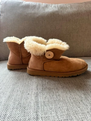 UGG mini bailey button  - Jag säljer ett par UGG mini bailey button i färgen kastanj i storlek 36. Finns en fläck på framsidan av den högra skon men har ej testat att få bort