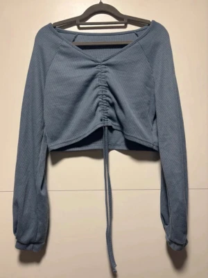 Blå croppad topp med rynk framtill - Trendig blå croppad topp med långa ärmar och rynkdetalj framtill som justeras med snören. Tillverkad i ribbat tyg för en cool och avslappnad look. Perfekt för att styla med höga jeans eller kjol.