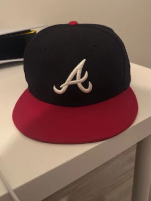 Atlanta Braves svart/röd keps New Era 7 1/4 - Snygg svart keps från New Era med röd skärm och broderad vit 'A' framtill, klassisk Atlanta Braves-design. Baksidan har MLB-logga. Perfekt för dig som gillar streetwear och sportig stil.