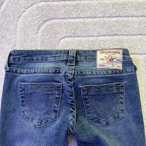 True Religion blå bootcut jeans - Lågmidjade True religion jeans blå med ljusrosa sömmar, har ett litet hål på knät men inget som syns.storlek 24 passar xs-s