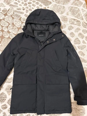Svart parkas från Peak Performance - Pris online 6000kr denna jacka är en snygg svart parkas från Peak Performance med huva, dragkedja och tryckknappar framtill. Jackan har flera praktiska fickor och en stilren design. Perfekt för kalla dagar tack vare det vadderade materialet och den långa modellen.