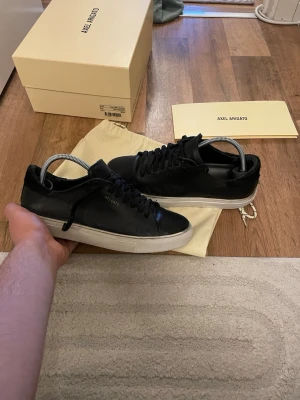 Svarta Axel Arigato sneakers i läder - Stilrena svarta sneakers från Axel Arigato i slätt läder med vit sula och diskret guldfärgad logga på sidan. Klassisk låg modell med svarta skosnören och minimalistisk design. Perfekta för dig som gillar clean och modern stil.