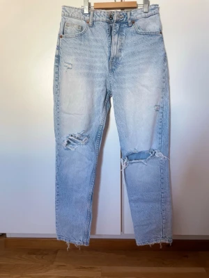 Ljusa slitna högmidjade ”mom” jeans - Säljer ett par ljusblå mom jeans med hög midja och raka ben. Jeansen har slitningar och hål på båda benen för en trendig, avslappnad look. Klassisk femficksmodell och dragkedja med knapp. Perfekta för en cool och avslappnad stil.
