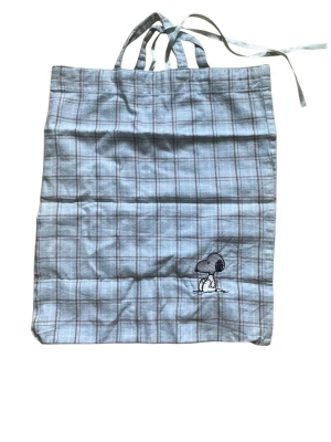 Rutigt tygpåse med Snoopy-motiv - Ljusblå tygpåse med rutigt mönster och broderad Snoopy-figur i hörnet. Påsen har långa band för att knyta och är tillverkad i mjukt bomullstyg. Vintage från tidigt 00 tal eller sent 90-tal.