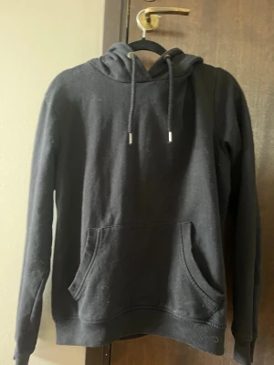Svart hoodie med magficka - En klassisk svart hoodie med huva och dragsnören. Hoodien har en stor magficka framtill och ribbade muddar vid ärmslut och nederkant. Materialet känns mjukt och bekvämt. Köpt på Ullared. Säljer för att den inte används. Storlek 36-38.