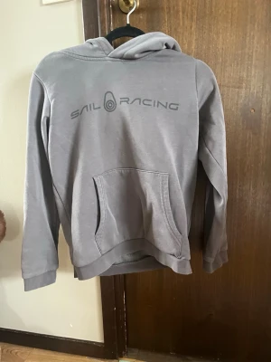 Grå hoodie från Sail Racing - Snygg grå hoodie från Sail Racing med tryckt logga på bröstet. Klassisk känguruficka framtill och huva. Mjuk bomullskänsla och ribbade muddar vid ärmslut och nederkant. Säljer för att den är för liten. Storlek 150.