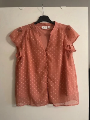 Rosa blus från Vila - Söt rosa blus från Vila med små prickar i tyget och knappar framtill. Blusen har korta ärmar med lätt puff och är tillverkad i ett tunt, transparent material som ger en luftig känsla. Perfekt för en trendig och avslappnad look.