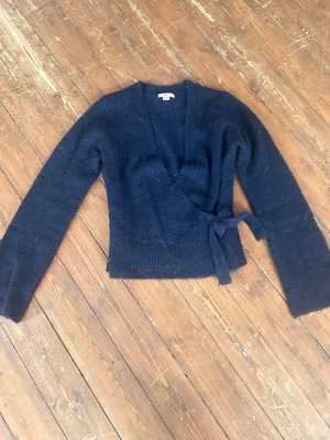 Mörkblå omlottstickad tröja - Mysig mörkblå stickad tröja i omlottmodell med knytband i midjan. Tröjan har långa ärmar och ribbstickade muddar vid ärmslut och nederkant. Perfekt för lager på lager och en avslappnad men snygg look. 