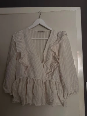 Vit volangblus från ONLY - Söt vit blus från ONLY med tunna vertikala ränder och volangdetaljer längs framstycket och axlarna. Blusen har omlott-skärning, resår i midjan och långa ärmar med lätt puff. 