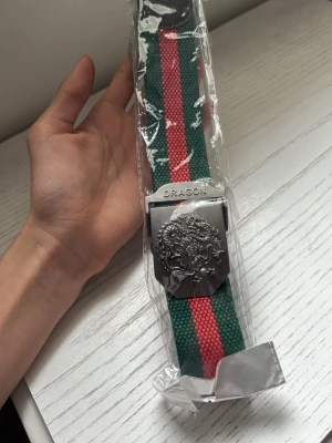 Randigt bälte från Dragon - Säljer ett randigt bälte från Dragon med gröna och röda färger. Bältet har en metallspänne med drakmotiv och texten 'DRAGON' ingraverad. Materialet är syntet och spännet är silverfärgat. Perfekt för att ge din outfit en cool touch.