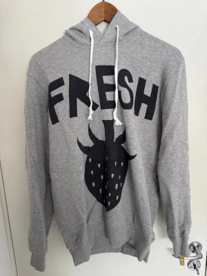 Grå Comme des Garcons hoodie - Grå hoodie med svart tryck där det står 'FRESH' och en stor jordgubbe på framsidan. Tröjan har huva med vita snören och ribbade muddar vid ärmar och nederkant. Tillverkad i mjukt sweatshirtmaterial, perfekt för en avslappnad stil.