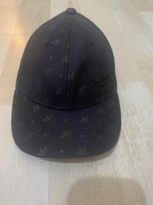 Svart Adidas keps med diskret mönster - Snygg svart keps från Adidas med broderad logga framtill och diskret mönster av små Adidas Trefoil-symboler över hela kepsen. Klassisk böjd skärm och justerbar passform. Tillverkad i slitstarkt tyg som passar perfekt till streetwear-stilen.