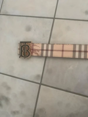 Burberry bälte med TB-spänne - Säljer ett snyggt Burberry bälte med klassiskt rutmönster i beige, brunt och svart. Bältet har ett guldigt TB-spänne som verkligen sticker ut. Materialet är läder och canvas, perfekt för att lyfta din outfit med en ikonisk accessoar.