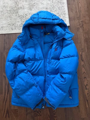 Blå dunjacka från Polo Ralph Lauren - Snygg blå dunjacka från Polo Ralph Lauren med huva och dragkedja framtill. Jackan har broderad logga på bröstet, flera fickor med dragkedja och en puffig, varm design perfekt för kalla dagar. Klassisk modell med justerbar huva och detaljer i metall.