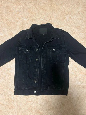 Svart jeansjacka med silverknappar - Snygg svart jeansjacka med klassisk krage och silverfärgade knappar. Två bröstfickor med lock och knapp framtill. Rak passform och stilrena sömmar ger en clean look. Perfekt att slänga över en hoodie eller t-shirt för en avslappnad vibe.