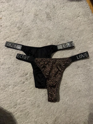 Love Secret stringtrosor med glitterband - Två stringtrosor från Love Secret, en i svart och en med leopardmönster. Båda har breda glitterband med texten 'LOVE SECRET' i silver och svart. Materialet är mjukt och stretchigt, perfekt för dig som gillar att sticka ut med detaljer.