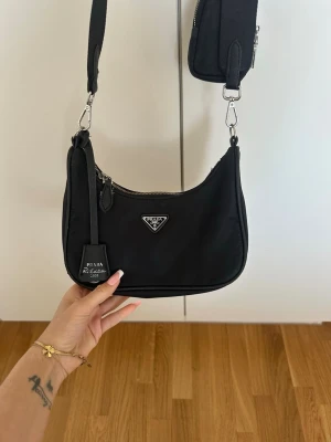Svart nylonhandväska från Prada - Snygg svart handväska från Prada i nylon med silverfärgade detaljer och avtagbar axelrem. Defekter förekommer i form av att silvret blivit mörkare i dragkedja samt en hake, går att byta ut hos väsk reparatör. Även har innertyget blivit utklippt då det gick sönder, därav priset. 