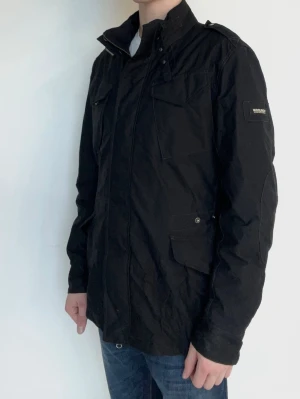 Svart vindjacka från Woolrich - Stilren svart vindjacka från Woolrich med hög krage, fyra fickor framtill och diskret logotyp på ärmen. Jackan har en rak passform och är tillverkad i ett slitstarkt material som skyddar mot vind. Perfekt för dig som vill ha en clean och funktionell look. Modellen är ca 182, 70 kg.