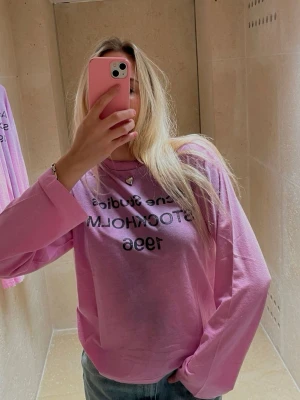 Ljusrosa långärmad topp Acne Studios - Ljusrosa långärmad topp från Acne Studios med svart tryck 'Acne Studios STOCKHOLM 1996' på bröstet. Klassisk rund halsringning och relaxed fit. Perfekt för dig som gillar stilrena och trendiga plagg med en touch av streetwear.