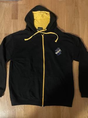 AIK Hoodie Large (L)  - Svart AIK huvtröja med dragkedja och luva i mycket fint skick. Tröjan har en gul dragkedja och gula snören i luvan. På bröstet finns ett broderat AIK-märke i blått och gult. Insidan av luvan är också gul. Tröjan har två fickor framtill. Baksidan är helsvart.   Kan även hämtas i Vallentuna eller Östra station om man vill slippa fraktkostnaden.