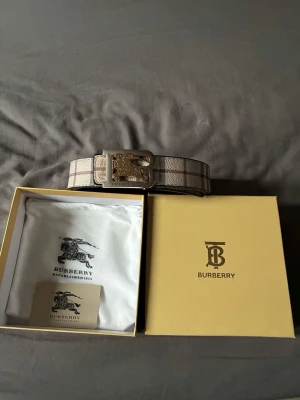 Designinspirerat rutigt bälte med gulddetaljer - Stilrent bälte inspirerat av Burberry i klassiskt rutigt mönster med beige, bruna och röda toner. Går vända på och få en helt annan stil på det. Bältet har ett exklusivt spänne i guld med Burberry-inspirerad logga och ikoniska ryttare. Perfekt accessoar för att lyfta din outfit.