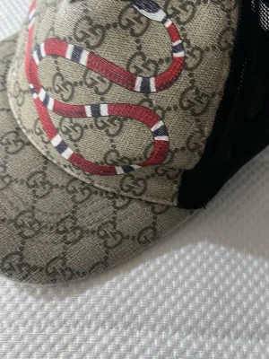 Gucci keps med orm och GG-mönster - Säljer en beige Gucci keps med klassiskt GG-monogram och en röd, vit och svart orm på framsidan. Kepsen har svart mesh på baksidan för extra ventilation och justerbar passform. Perfekt för dig som vill sticka ut med en ikonisk accessoar.
