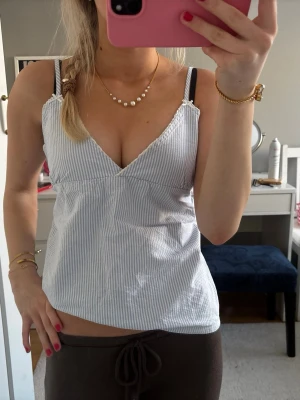 Randig vit och blå linnetopp - Säljer en snygg randig linnetopp i vitt och ljusblått med v-ringning och tunna axelband. Toppen har en enkel och fräsch design som passar perfekt till jeans eller shorts. Materialet känns lätt och somrigt.