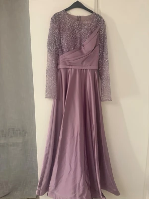 Ljuslila långklänning med glitterärmar - Elegant långklänning i ljuslila med glittriga, långärmade spetsärmar och asymmetrisk drapering över bysten. Kjolen är vid och faller snyggt. Passar bra till bal eller andra festliga tillfällen