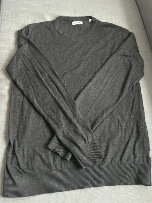 Mörkgrå stickad tröja Jack & Jones - Mörkgrå stickad tröja från Jack & Jones med rund halsringning och långa ärmar. Tröjan har ribbade muddar vid ärmslut och nederkant, vilket ger en snygg och enkel look. Perfekt för lager på lager och passar till det mesta.