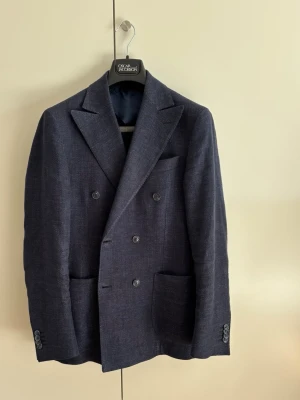 Oscar Jacobson Erik Double-Breasted Blazer - Ytterst sparsamt använd dubbelknäppt kavaj från Oscar Jacobson som dessvärre blivit för liten. Inga fläckar eller skador. Endast lätt skrynklig efter förvaring och blir i princip som ny efter pressning eller ångning.  Modellen är Erik, som är med smal passform och lite spetsigare slag. Tillverkad i en strukturväv av lin och bomull, signerad den italienska vävproducenten Lanificio Angelico. Kavajen är halvfodrad, tre innefickor, varav en pennficka.  Nypris: 5299 SEK  Storlek: 46 (S)