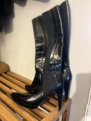Svarta höga stövlar Novita Scorett - Säljer ett par svarta höga stövlar med spetsig tå och smal klack. Stövlarna har en snygg glansig finish i skinnimitation och paneler i stretchigt tyg längs skaftet för en tight passform. Perfekta för att lyfta vilken outfit som helst.