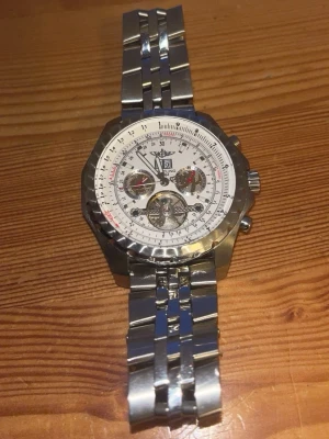 Silverfärgad Breitling klocka - Snygg och detaljerad klocka från Breitling med silverfärgad länkarmband i metall. Urtavlan är vit med flera små ur och tydliga markeringar, samt logga och datumvisning. Baksidan har ingraverade bilar och modellnamn. Perfekt accessoar för dig som gillar stilrena detaljer.