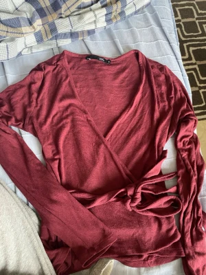 Vinröd omlottopp från 157 - Snygg vinröd omlottopp från 157 med långa ärmar och knyt i midjan. Toppen har en djup v-ringning och är tillverkad i ett mjukt material som känns skönt mot huden. Perfekt för dig som gillar en enkel men trendig stil.
