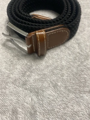 Svart flätat bälte med bruna detaljer - Snyggt svart flätat bälte med bruna skinn-detaljer och silverfärgat spänne. Perfekt för att ge din outfit en stilren touch. Bältet har en flexibel design och passar till många olika looks.