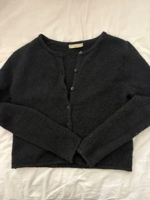 Svart ullkofta från Gina Tricot  - Mysig svart kofta från Gina Tricot ullblandning. Kortare modell med rund halsringning och knäppning framtill med svarta knappar. Långärmad och enkel design som passar perfekt till de flesta sammanhang! 