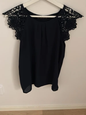 marinblå blus med spets från Zara Basic - Snygg marinblå blus från Zara Basic med vackra spetsdetaljer på axlarna. Blusen har rund halsringning och är ärmlös, vilket ger en elegant och stilren look. Perfekt att matcha med både jeans och kjol för en trendig outfit.