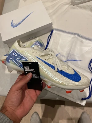 Nike Mercurial fotbollsskor  - Snygga Nike Mercurial fotbollsskor i skimrande vitt med blå detaljer och stor blå Swoosh på sidan. Skorna har orange dobbar och är tillverkade i ett lätt syntetmaterial för bästa grepp och fart på planen. Perfekta för dig som vill sticka ut på matchen.