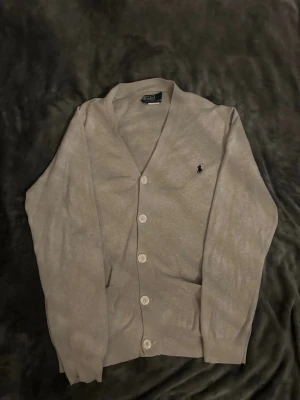 Beige cardigan från Polo Ralph Lauren - Klassisk beige cardigan från Polo Ralph Lauren med v-ringning och fem vita knappar framtill. Tröjan har lång ärm och den ikoniska mörkblå logotypen broderad på bröstet. Perfekt att slänga över en t-shirt för en clean och stilren look.