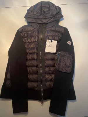 Svart dunjacka med huva från Moncler - Snygg svart dunjacka från Moncler med stickade ärmar och vadderad front. Jackan har huva, dragkedja framtill och en praktisk ficka med dragkedja på ena ärmen. Moncler-logga på ärmen och ribbade muddar för extra komfort.