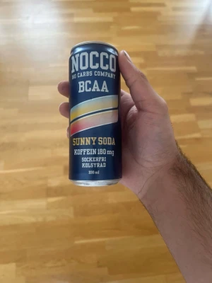 NOCCO BCAA Sunny Soda 330 ml burk - NOCCO BCAA Sunny Soda är en sockerfri och kolsyrad dryck med 180 mg koffein i en blå metallburk på 330 ml. Burken har färgglada detaljer och tydlig märkning. Perfekt för dig som vill ha en energiboost utan socker.