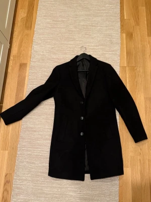 Svart klassisk ullkappa storleken S - Stilren svart ullkappa med tre knappar framtill och två fickor. Kappan har en rak passform, slät yta och klassisk krage. Inte överdrivet lång. Storlek S