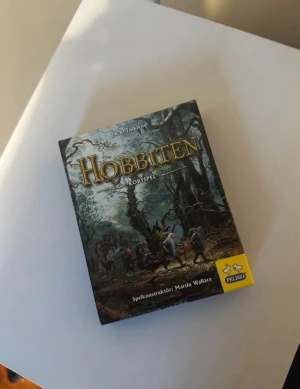 Hobbiten kortspel från Peliko - Säljer kortspelet Hobbiten, baserat på J.R.R. Tolkiens klassiker. Spelet har ett fantasytema med illustrationer av karaktärer som Bilbo, Thorin och Gandalf. Lådan är svart med färgstarka motiv och innehåller 65 kort samt regler. Perfekt för spelkvällar med vänner.