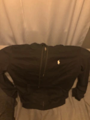 Svart ziptröja från Polo Ralph Lauren - Svart ziptröja från Polo Ralph Lauren med klassisk vit broderad logga på bröstet. Tröjan har dragkedja framtill, ribbade muddar och en enkel, stilren design. Perfekt för en avslappnad och snygg look.